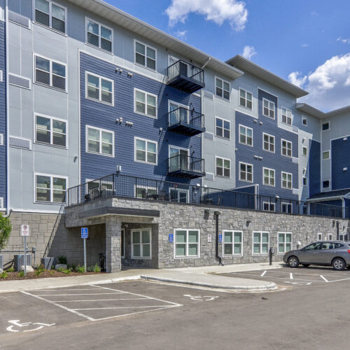 Modern multi-story apartment building with blue and gray exterior, balconies, parked cars, and designated handicap parking spaces in front—welcome to Flats at Neill Park, stylish apartments in Burnsville MN.