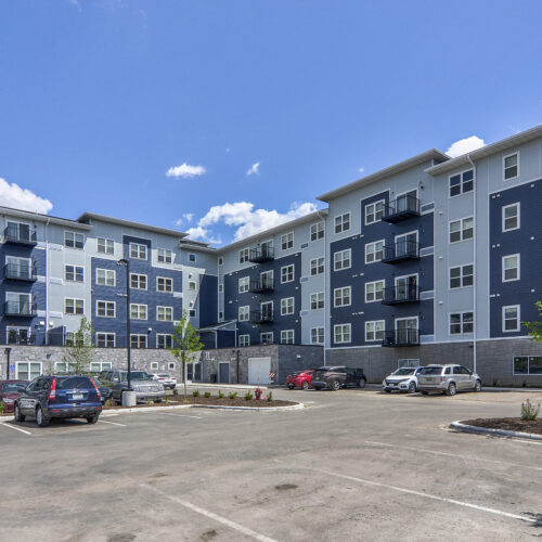 A modern, multi-story apartment building with balconies and a parking lot in front, under a blue sky with scattered clouds—the perfect home at Flats at Neill Park, offering some of the finest apartments in Burnsville MN.