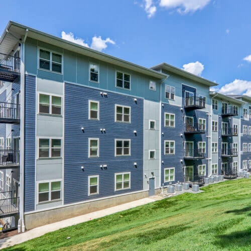 Four-story modern apartment building—Flats at Neill Park—with gray siding, multiple windows, and outdoor balconies, set on a grassy slope under a partly cloudy sky. Discover stylish apartments in Burnsville MN with scenic views.