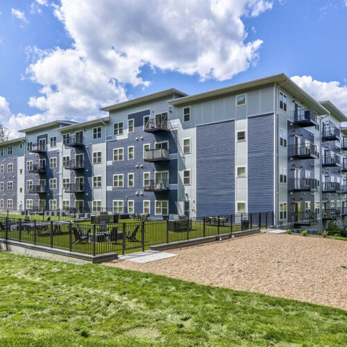 Flats at Neill Park is a four-story modern apartment building with balconies, fenced courtyard, and an adjacent sand area, surrounded by green lawn and trees under a partly cloudy sky—perfect for those seeking apartments in Burnsville MN.