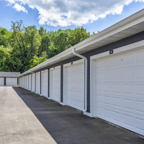 Row of closed white garage doors attached to a building at Flats at Neill Park, with more free-standing garages and trees in the background under a partly cloudy sky—ideal for those seeking apartments in Burnsville MN.