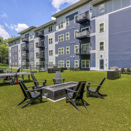 Outdoor communal area with artificial grass, black chairs around a fire pit, ping pong table, and the modern Flats at Neill Park apartments in Burnsville MN as the backdrop.