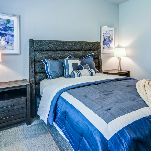 A neatly made bed with blue and white bedding, two bedside tables with lamps, framed artwork on the walls, and an open closet in a modern bedroom at Flats at Neill Park—stylish apartments in Burnsville MN.