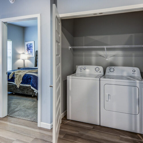 A laundry area with a washer and dryer inside a closet sits next to a bedroom with a bed, nightstand, and lamp—perfect for comfortable living at Flats at Neill Park, one of the top apartments in Burnsville MN.
