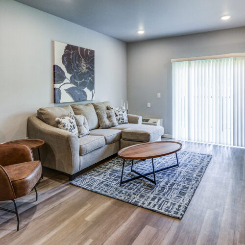 Modern living room at Flats at Neill Park with a beige sofa, brown armchair, wooden coffee table, patterned rug, and sliding glass doors with vertical blinds—an inviting space in apartments in Burnsville MN.