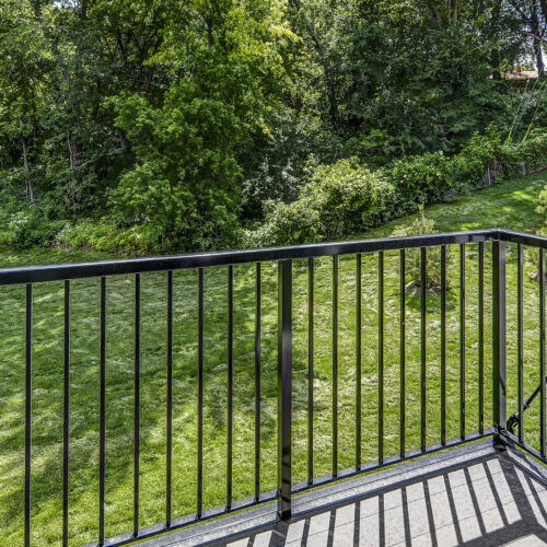 View from a balcony with black metal railing overlooking a green grassy yard and dense trees in the background at Flats at Neill Park, one of the premier apartments in Burnsville MN.