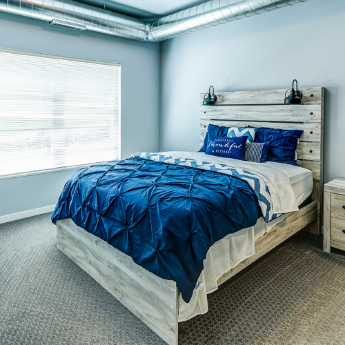 Bedroom at Flats at Neill Park with a wooden bed frame, blue and white bedding, matching nightstand, large window, and exposed ductwork—perfect for those seeking stylish apartments in Burnsville MN.