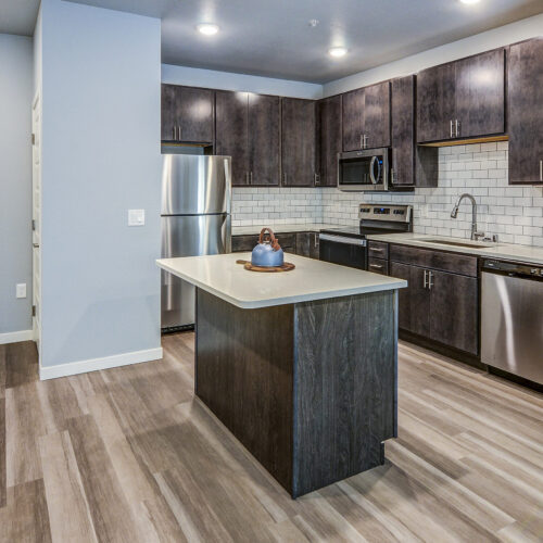 Modern apartment kitchen at Flats at Neill Park with dark wood cabinets, stainless steel appliances, white subway tile backsplash, and a central island on light wood flooring—premium apartments in Burnsville MN.