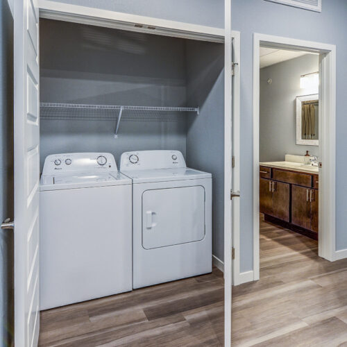 A laundry closet with a washing machine and dryer, open double doors, shelf above appliances; adjacent to a bathroom and entry door at Flats at Neill Park, featuring wood-style flooring throughout these apartments in Burnsville MN.