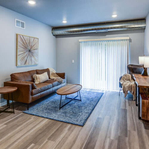 Modern living room at Flats at Neill Park with a brown leather sofa, round wooden coffee table, blue rug, wooden console table, and sliding glass doors with vertical blinds. Bedroom visible through open door—stylish apartments in Burnsville MN.