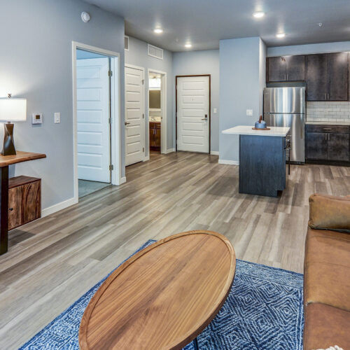 Modern open-plan apartment with wood flooring, stainless steel appliances, brown sofa, round coffee table, and a console table with a circular mirror—perfect for those seeking stylish apartments in Burnsville MN.