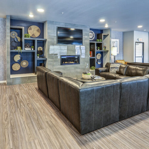 Modern lounge at Flats at Neill Park with a large black sectional sofa, built-in fireplace, wall-mounted TV, decorative shelving, leather chair, and wood-style flooring—stylish living in top apartments in Burnsville MN.