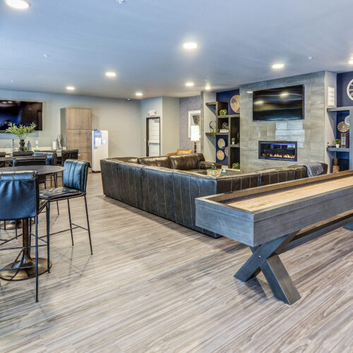 Modern communal lounge at Flats at Neill Park features leather seating, a wall-mounted TV, fireplace, shuffleboard table, dining tables, and open shelving—perfect for relaxing or socializing in these contemporary apartments in Burnsville MN.