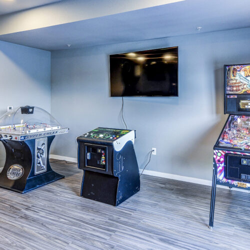 Three arcade machines, including a bubble hockey table, driving game, and pinball machine, stand against a gray wall beneath a TV in the game room at Flats at Neill Park—modern apartments in Burnsville MN with great amenities.