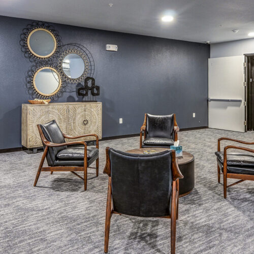 A modern lounge area at Flats at Neill Park features four black leather chairs around a round table, a patterned sideboard, decorative mirrors, and a dark blue accent wall—perfect for relaxing in these stylish apartments in Burnsville MN.