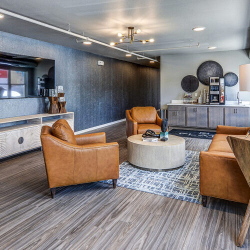 Modern lounge at Flats at Neill Park features brown leather chairs, a round coffee table, TV on the wall, sideboard, and floor lamp atop wood flooring with industrial ceiling details—perfect for apartments in Burnsville MN.