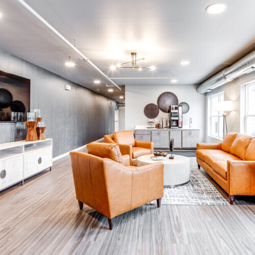 Modern lounge area at Flats at Neill Park featuring tan leather sofas, a round ottoman, wall-mounted TV, wooden sideboard, decorative vases, and large windows filling these apartments in Burnsville MN with natural light.