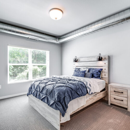 Modern bedroom at Flats at Neill Park with exposed ductwork, a large window, blue bedding on the bed, and a wooden nightstand on a carpeted floor—stylish comfort in apartments in Burnsville MN.