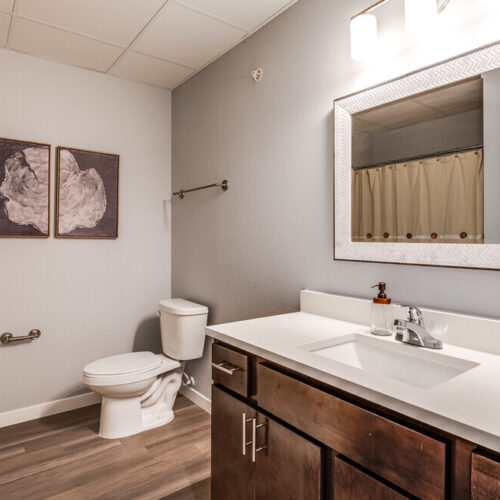 Modern bathroom at Flats at Neill Park features a toilet, large mirror above a sink with wooden vanity, two wall art pieces, towel rack, and a shower with a beige curtain—perfect for stylish apartments in Burnsville MN.