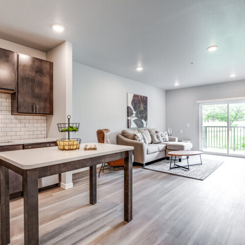 Modern apartment interior with an open kitchen and living area, wooden floors, brown cabinets, a sofa, large window, and a small balcony overlooking greenery—perfect for those seeking stylish apartments in Burnsville MN.