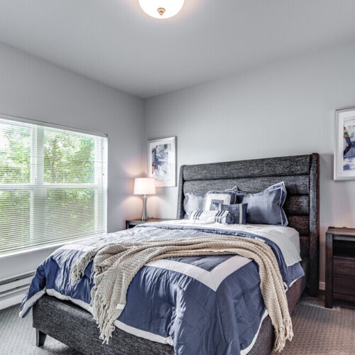 A neatly made bed with blue and white bedding in a bright bedroom at Flats at Neill Park, featuring two bedside tables with lamps, framed art, and a large window with blinds—perfect for apartments in Burnsville MN.