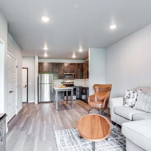 Modern open-concept living room and kitchen at Flats at Neill Park feature light wood floors, gray sofa, round wooden table, leather chair, and stainless steel appliances—perfect for those seeking stylish apartments in Burnsville MN.