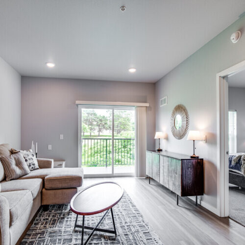 Modern living room with beige sofa, abstract art, round coffee table, and sliding glass doors leading to a balcony at Flats at Neill Park; adjacent bedroom visible through open doorway—perfect for stylish apartments in Burnsville MN.