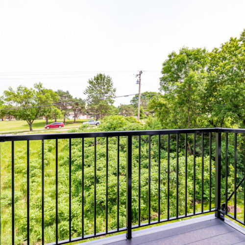 A black metal balcony at Flats at Neill Park overlooks a green, tree-filled landscape with a grassy area and a few cars and power lines in the background.