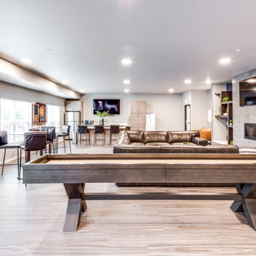 Spacious modern living area at Flats at Neill Park with a shuffleboard table in the foreground, lounge seating, two wall-mounted TVs, fireplace, dining area, and stylish decor on light wood flooring—perfect for those seeking apartments in Burnsville MN.