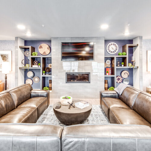 Modern living room at Flats at Neill Park with two brown leather sofas, a round coffee table, wall-mounted TV above a fireplace, built-in shelves with decor, and armchairs—ideal for those seeking stylish apartments in Burnsville MN.