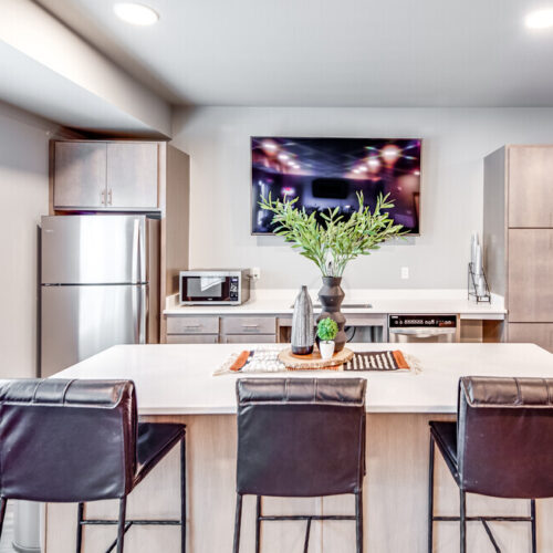 Modern kitchen with stainless steel appliances, a center island with three black chairs, a potted plant on the counter, and a wall-mounted TV—perfect for residents of Flats at Neill Park apartments in Burnsville MN.