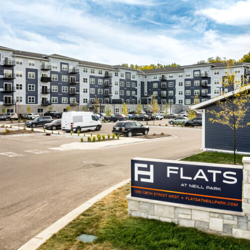 A modern apartment complex in Burnsville, MN, with a blue and white exterior, visible parking lot, parked cars, and a sign reading Flats at Neill Park.
