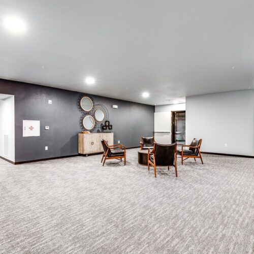 Modern hallway at Flats at Neill Park with gray carpet, white walls, exposed ductwork, and a seating area featuring four chairs and a sideboard with decorative mirrors—perfect for those seeking stylish apartments in Burnsville, MN.