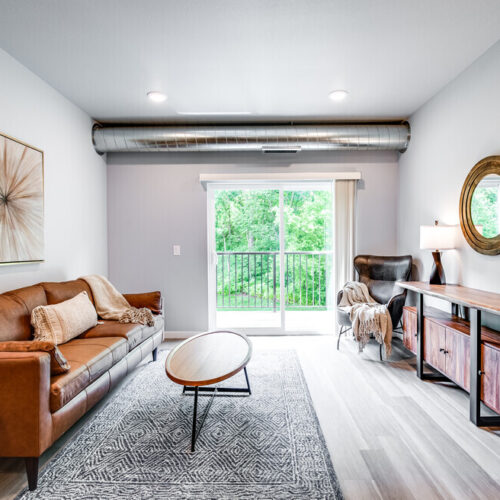 Modern living room at Flats at Neill Park features a brown leather sofa, oval coffee table, large window to a balcony, accent decor, and console table with lamps—perfect for those seeking stylish apartments in Burnsville MN.