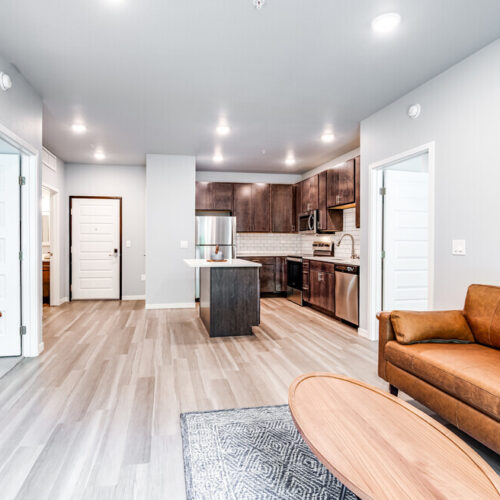 Modern open-concept living area at Flats at Neill Park with a brown leather sofa, wooden table, and kitchen featuring stainless steel appliances and dark wood cabinets—your stylish retreat among apartments in Burnsville MN.