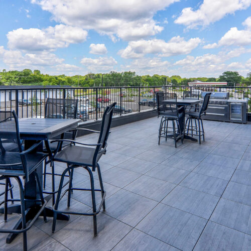 Outdoor rooftop patio with black tables and chairs, overlooking a parking lot and green trees under a partly cloudy sky.